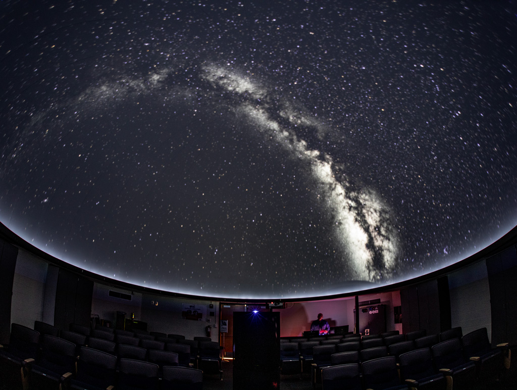 An image of the Milky Way arcs up along the dome of the Sidney Frohman Planetarium. The milky way is brilliant in black and white and floats amid a background of stars.