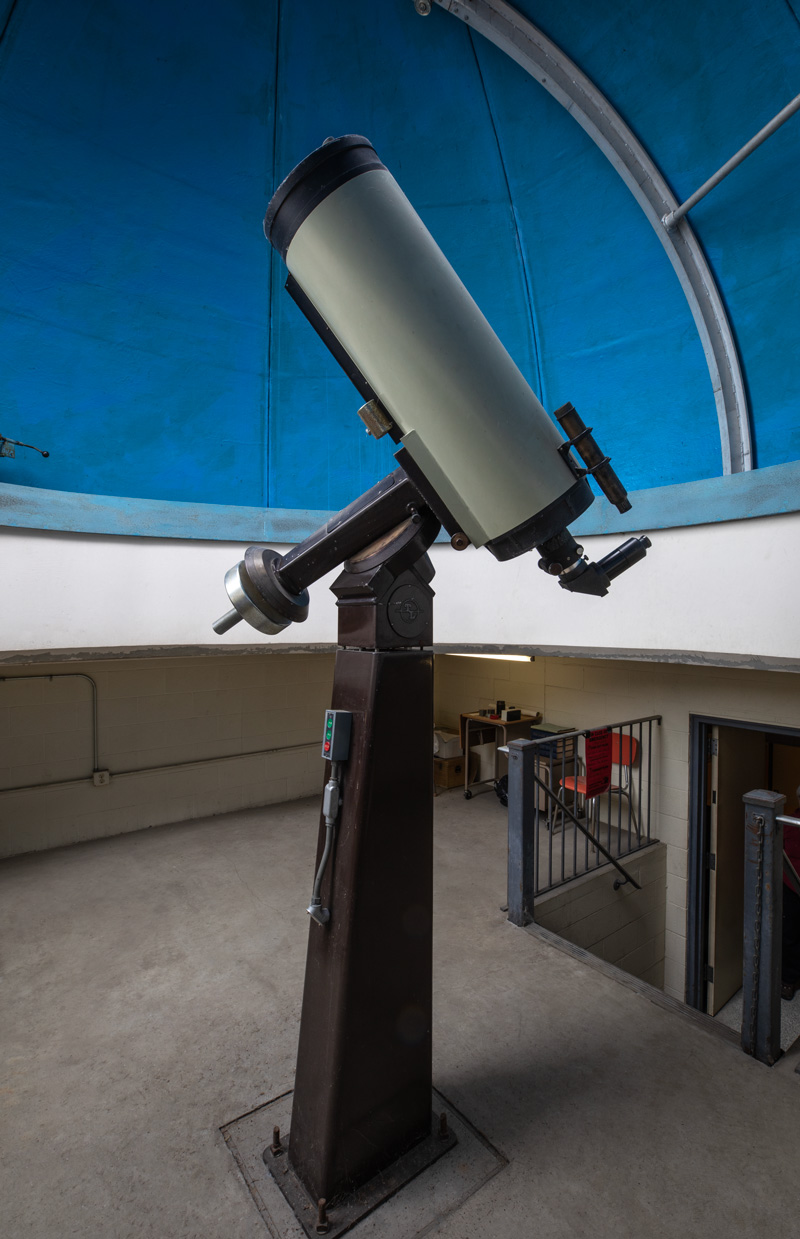 With the blue interior of its observatory dome as background, the white tube of the telescope points skyward. The instrument sits atop a brown steel pier and is counterbalanced by a sturdy weight.