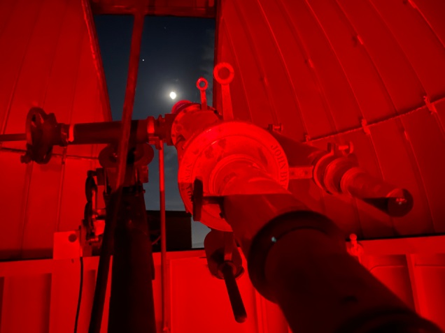 The ca. 1882 John Byrne telescope aimed at Moon for a night of observing. Photo by Dr. Ann Bragg, 2021. Under red light, the telescope points out the open aperture of its protective rooftop dome at Moon's glowing orb in a clear, dark sky.